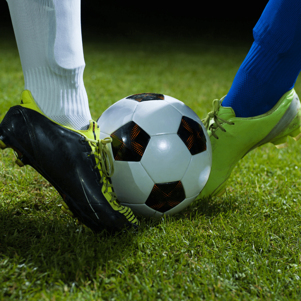 Two football boots meeting at the ball on a grass pitch at night