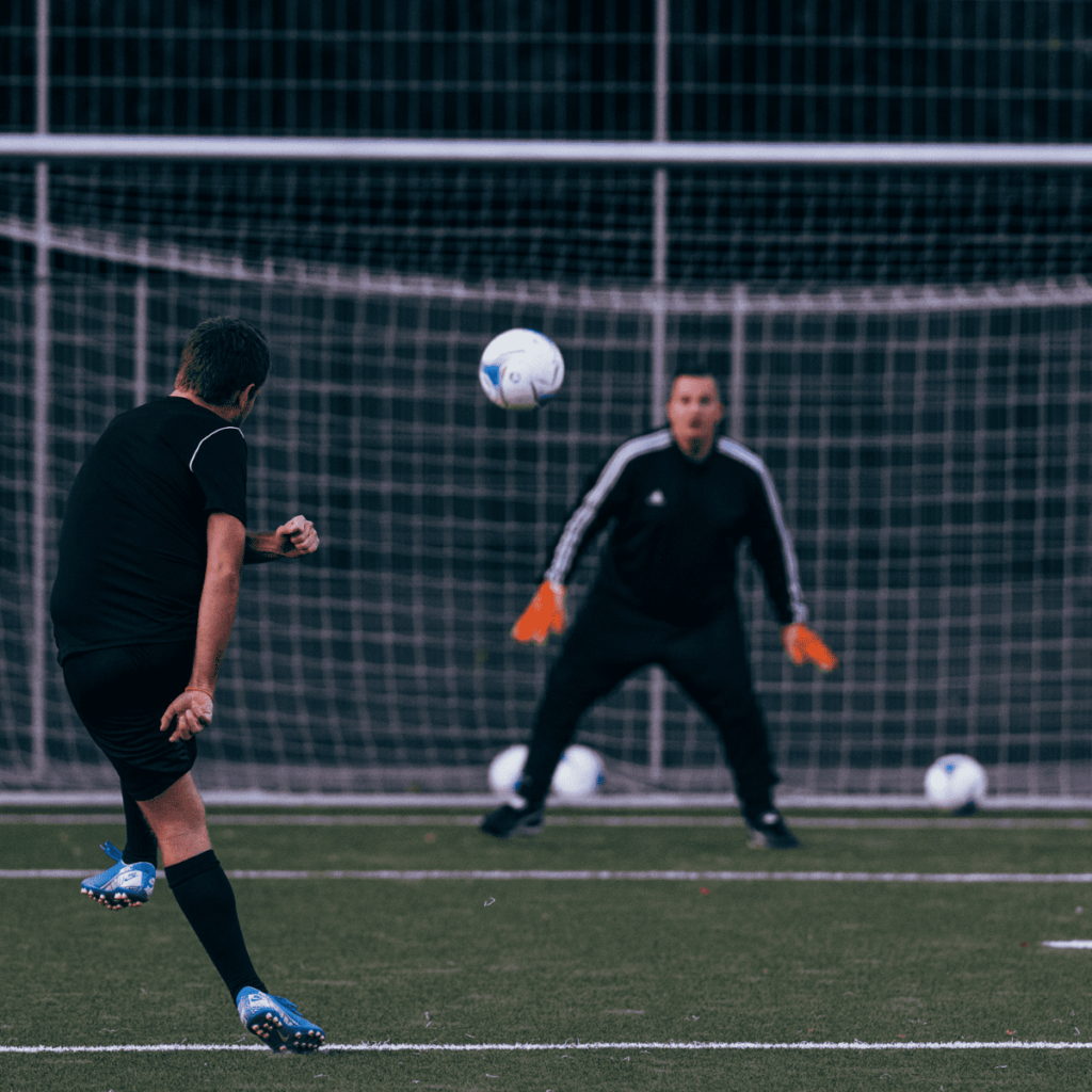 A player strikes the ball towards a goalkeeper waiting in front of the net on an artificial pitch
