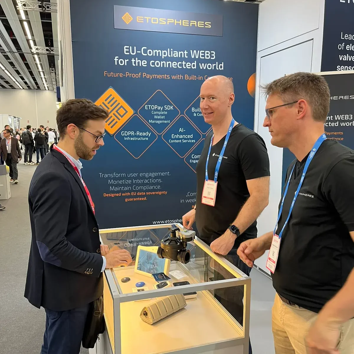 A visitor talks with two exhibitors at the Etospheres stand, a showcase with a valve and small devices in front of a banner on EU-compliant Web3 and GDPR-ready infrastructure