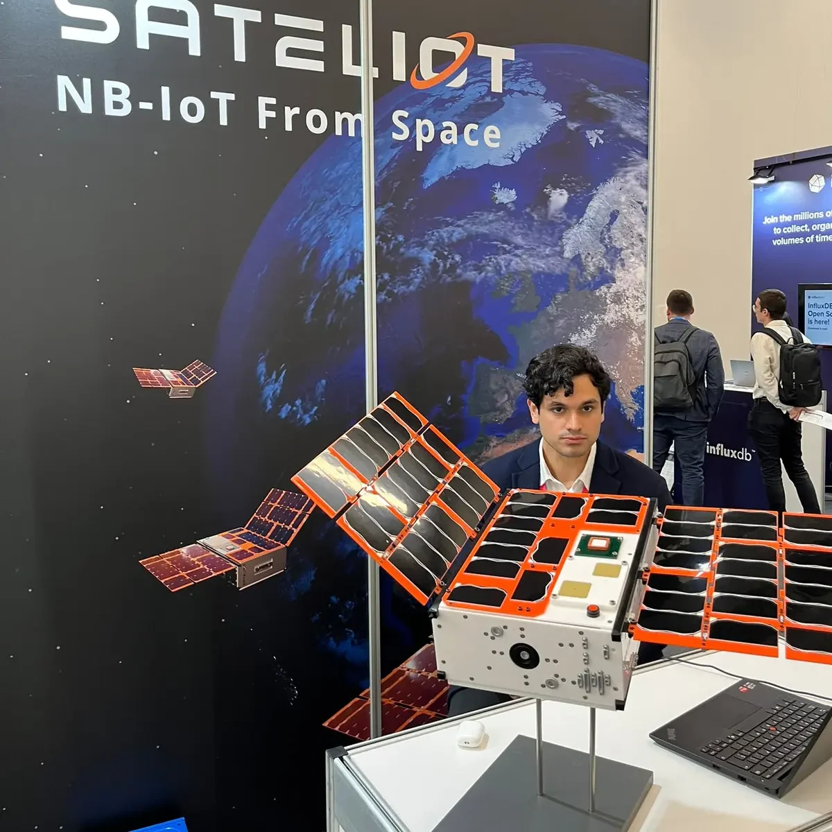 A satellite model with orange-framed solar panels on a stand, in front of a Sateliot backdrop that reads NB-IoT From Space