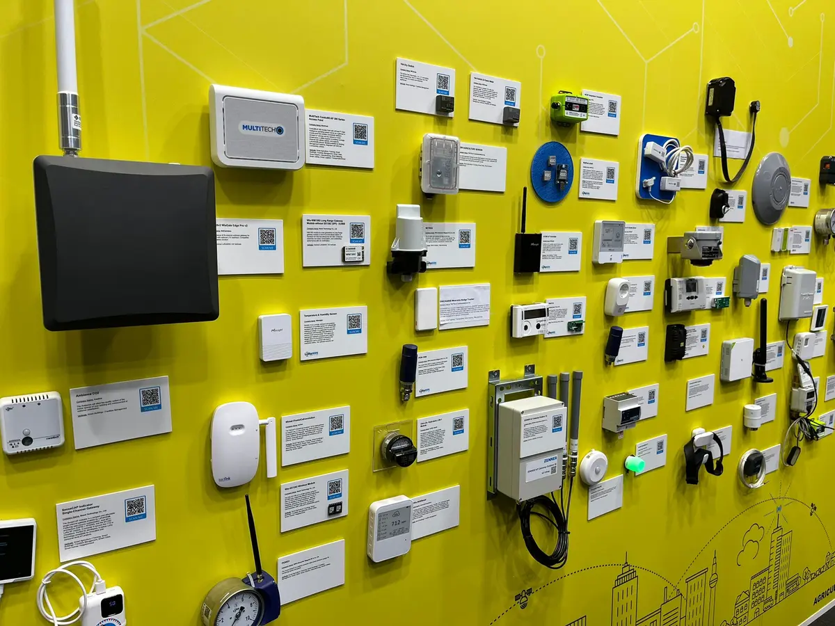 A yellow exhibition wall covered with gateways, sensors, meters and antennas, each with a label and a QR code