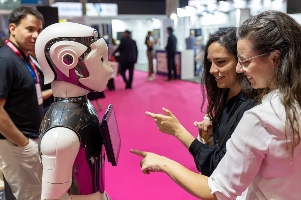 Two visitors smile and point at a white humanoid robot with a tablet on its chest, on the pink carpet of the exhibition floor