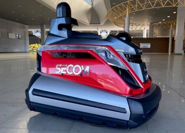 A red, silver and black Secom security robot standing on the polished floor of an airport terminal hall