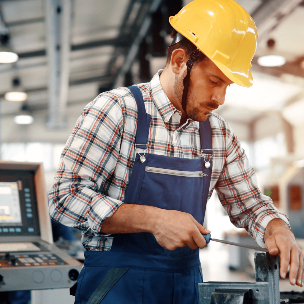 A factory worker in a yellow hard hat and overalls adjusts a part on a machine next to its control panel
