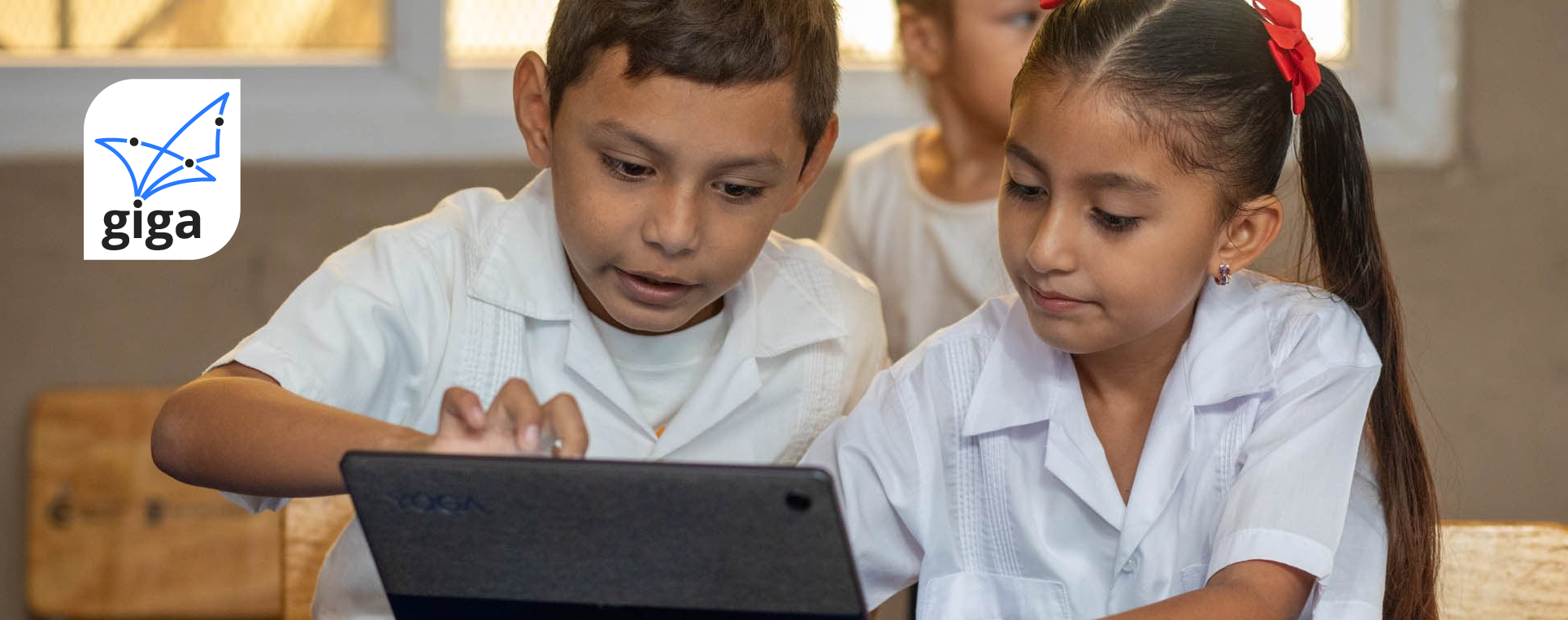 Two schoolchildren in white uniforms working together on a tablet in a classroom, with the Giga logo in the corner