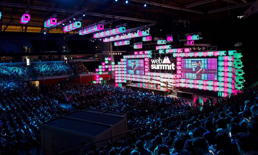 A packed arena facing the Web Summit main stage, framed by walls of pink and green light panels and two large screens showing a speaker