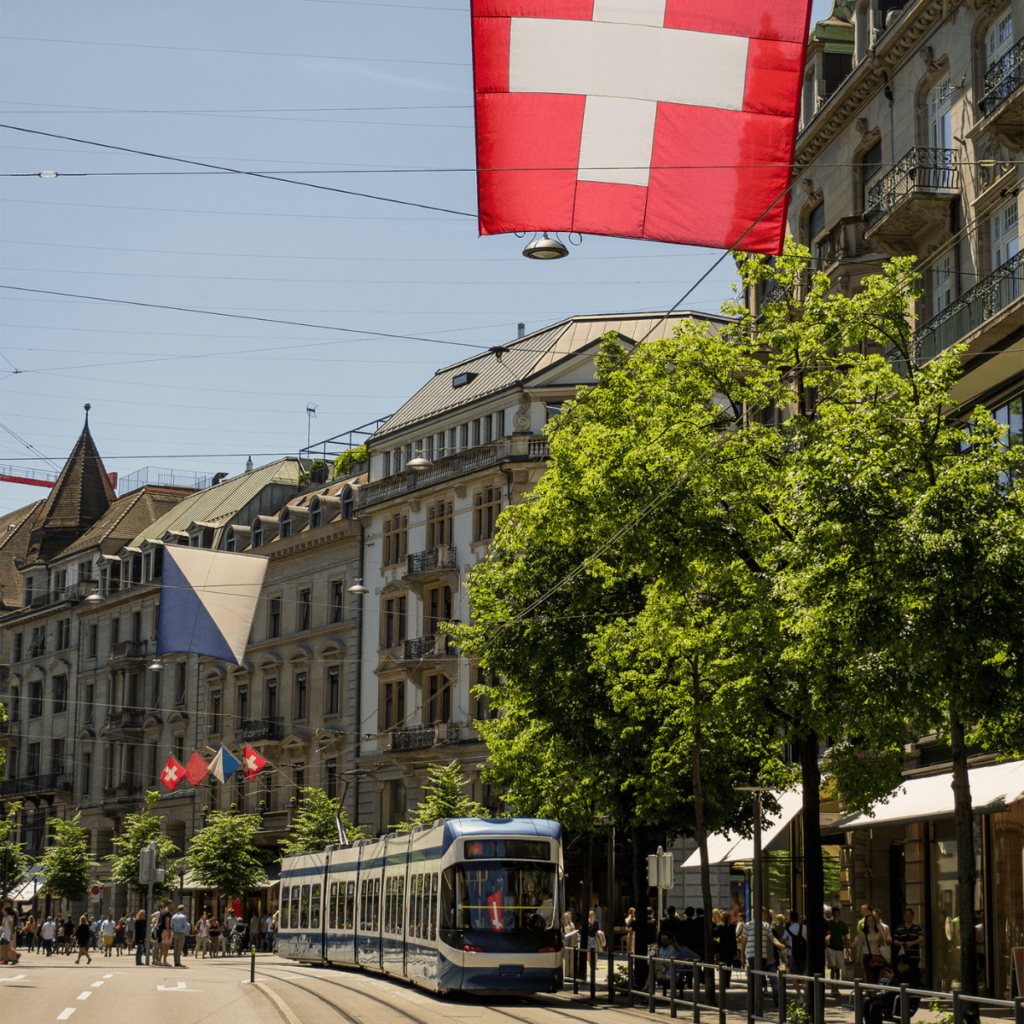 A blue and white tram runs along a tree-lined street of Zurich under a large Swiss flag hanging over the road