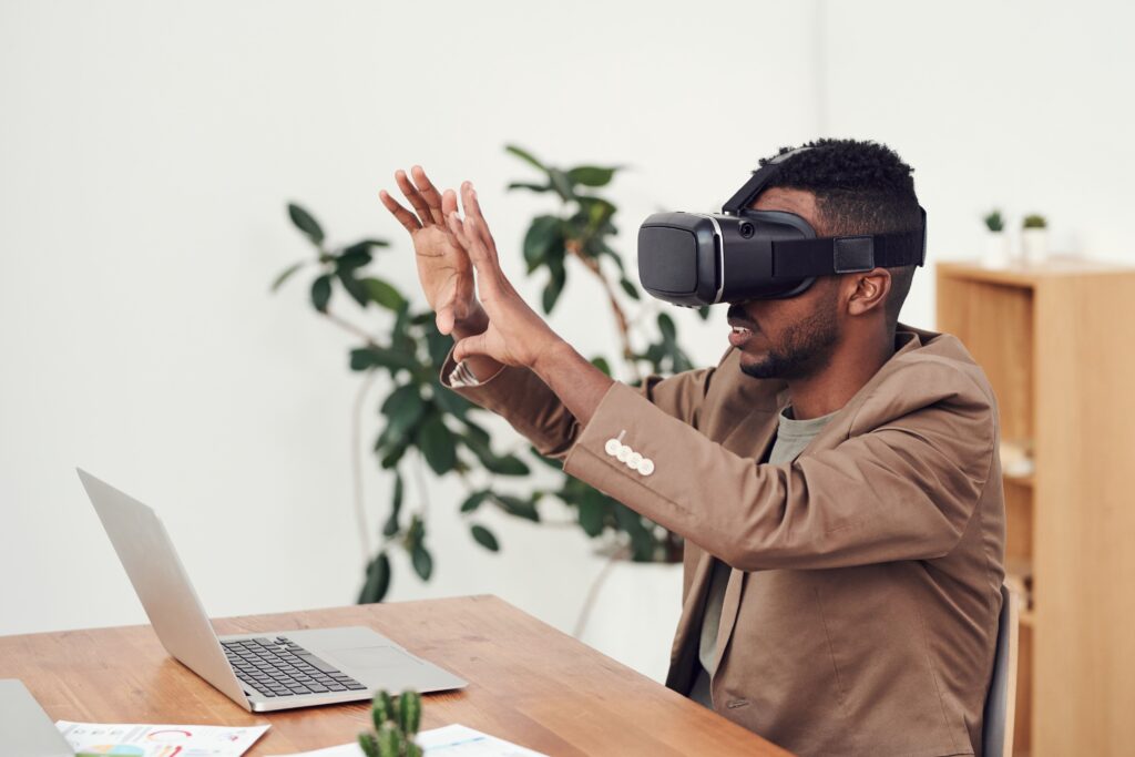 A man at a desk with a laptop wears a virtual reality headset and reaches out with both hands