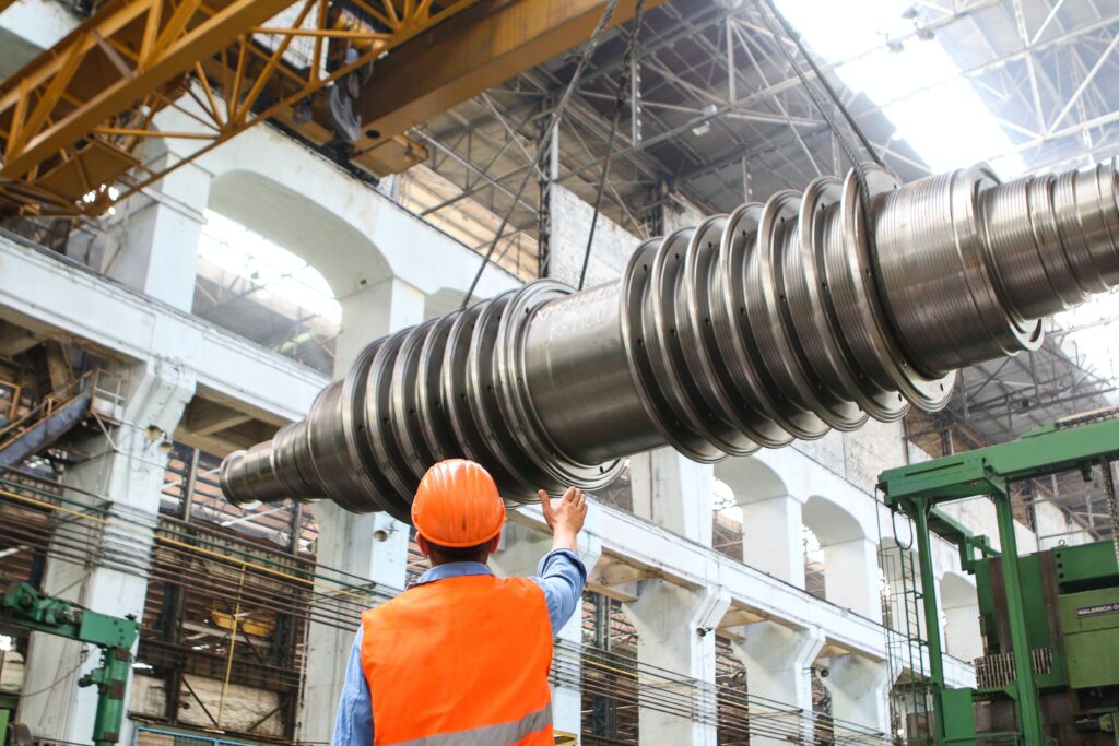 A worker in a hard hat and high-visibility vest guides a large turbine rotor hanging from an overhead crane in a factory hall